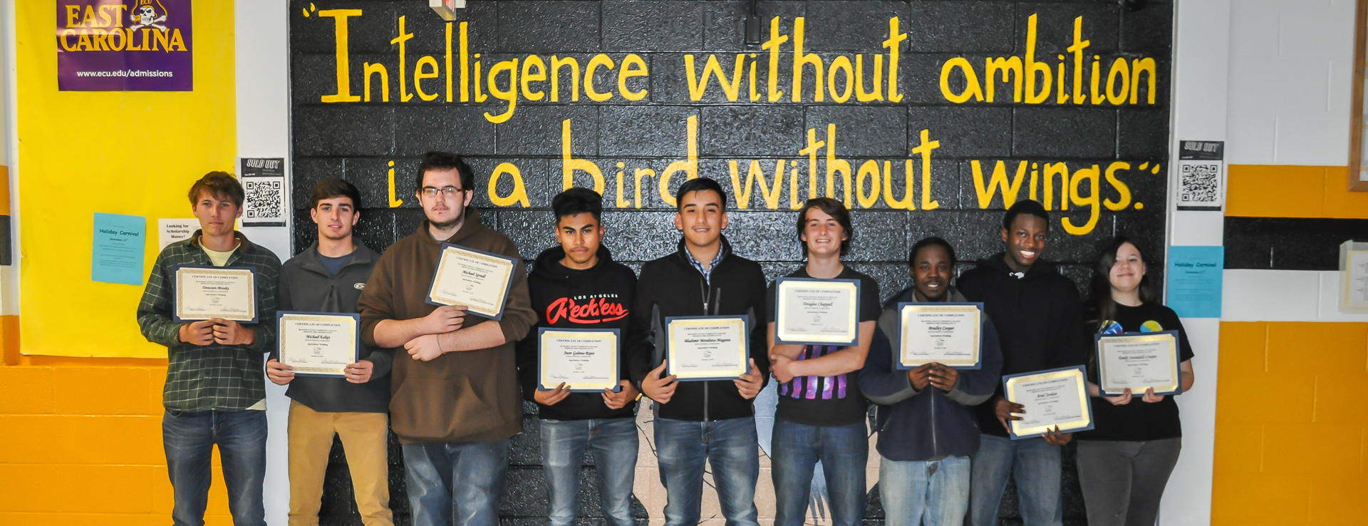 Nine students holding certificates in front of a wall reading 